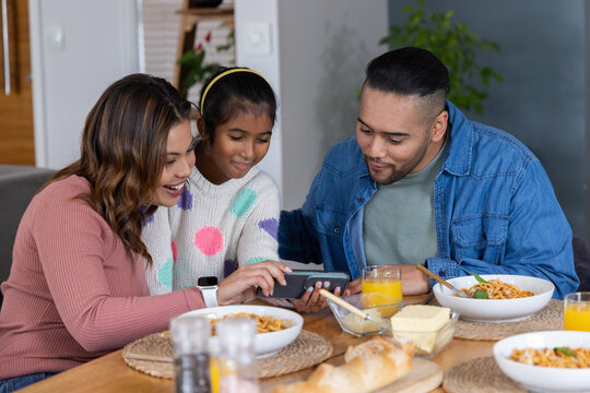 Parents with daughter leaning in, sharing phone at home dining table with pasta, bread, juice