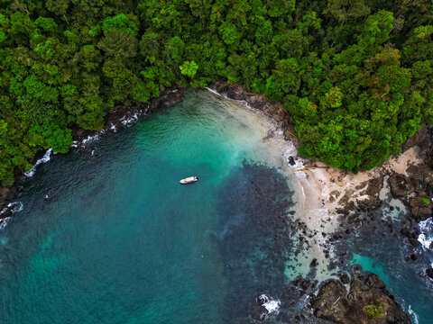 Aerial view of Bahia Solano featuring a small boat anchored in turquoise waters surrounded by dense tropical jungle and rocky shores in Mutis, Choco, Colombia.