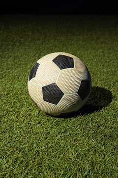 Textured black-and-white soccer ball is resting on short green grass, casting sharp shadow