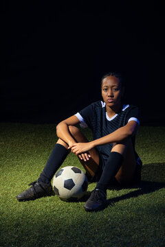 Female soccer player sitting on turf with black ball between feet wearing black kit and cleats