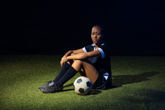 African American female soccer player sitting in light on turf wearing black kit with soccer ball
