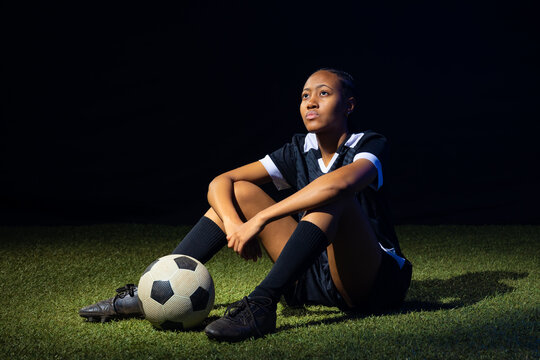 African American female soccer player sitting on turf at night, wearing dark jersey, holding ball