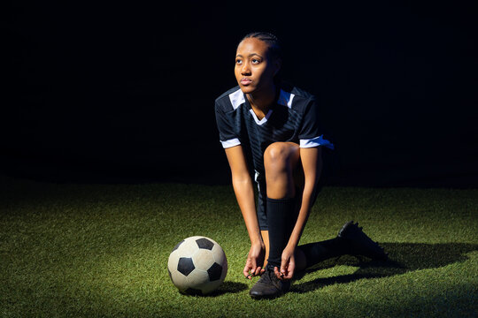 Adult female soccer player kneeling on turf tying cleat, wearing black-and-white kit, ball left