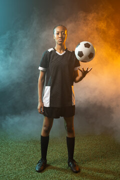 Woman soccer player standing on turf in studio balancing ball, wearing cleats, teal orange fog