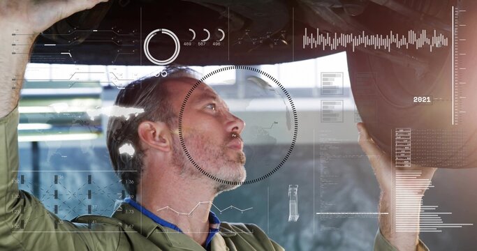 Working midlife mechanic in green coveralls blue-collared holding underbody parts in workshop, HUD