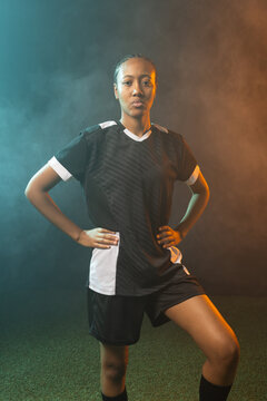 African woman posing on studio turf in black-white soccer kit, knee-high socks amid teal-amber haze