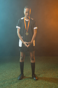 Female athlete standing on green turf in studio, wearing black soccer kit, holding gold medal