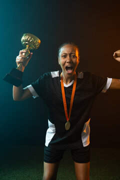 Female athlete wearing black-and-white jersey and shorts, holding gold trophy and medal on turf