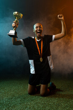 African American woman kneeling cheering holding gold trophy on turf in kit wearing medal, cleats