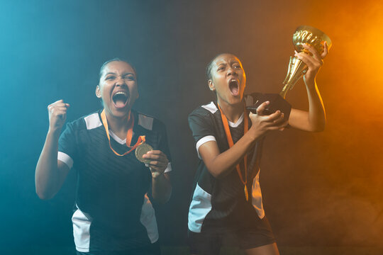 Two female athletes holding trophy, raising medal, wearing matching jerseys on studio set with haze