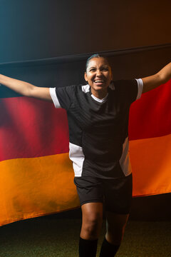 African female athlete holding black-red-gold tricolor flag on green turf, wearing black white kit