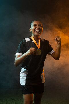 Female athlete celebrating on turf in black-white kit, holding gold medal amid blue-orange smoke