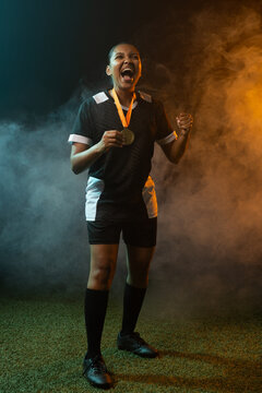 Female athlete shouting, holding gold medal on orange ribbon on turf amid smoke, wearing jersey
