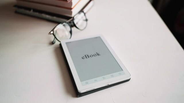 Electronic book reader displaying ebook text beside glasses and paper books on clean table. Concept of modern education, remote study, reading habit and convenient mobile knowledge access.
