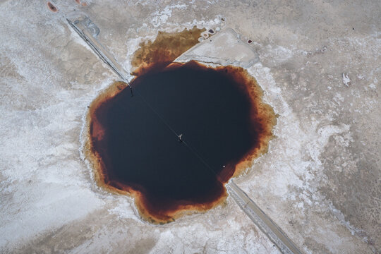 Aerial view of Searles Lake featuring a dark brine pool with reddish edges surrounded by white salt crust and a crossing utility line in San Bernardino County, California, United States.