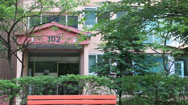 대한민국 서울의 여름철 아파트 단지 출입구 전경. 무성한 여름 나무들과 붉은색 출입구 지붕, 그리고 전경에 놓인 벤치가 어우러진 평화로운 한국의 주거 환경 자연 배경 이미지.