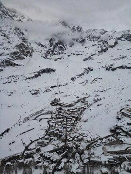 Aerial view of the snow-covered ski resort village nestled at the base of dramatic alpine peaks under a cloudy sky Breuil-Cervinia, Aosta Valley, Italy.