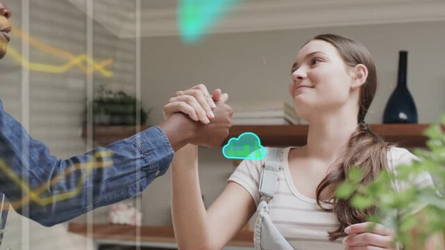 Two girls beside kitchen glass panel, raising and clasping hands and sending cloud overlay for tech