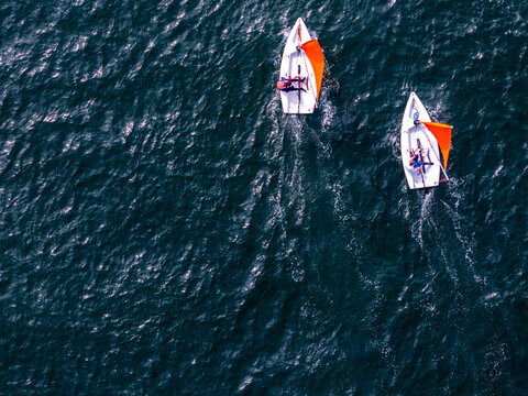 Aerial view of two small white sailboats with orange sails and people on board sailing across dark blue rippling water with white wakes.