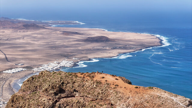 Aerial view of the volcanic coastline of El Golfo Lanzarote featuring white houses, rugged cliffs, and deep blue ocean waves under a clear sky El Golfo, Canarias, Spain.