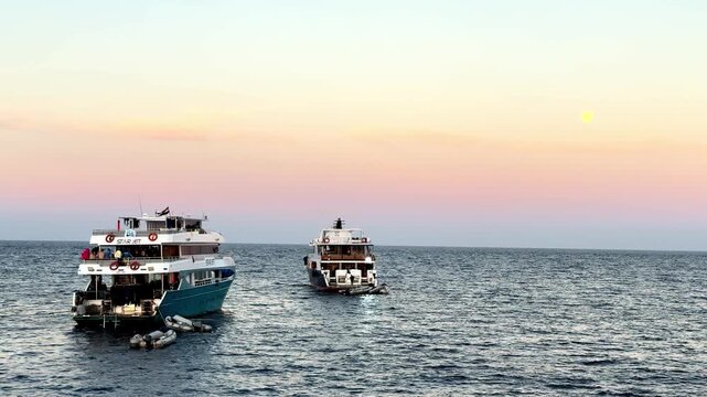 Two majestic white safari liveaboard cruise ships navigate deep blue tropical waters during a serene golden hour sunset with a glowing full moon in a clear pink sky from a low wide angle perspective