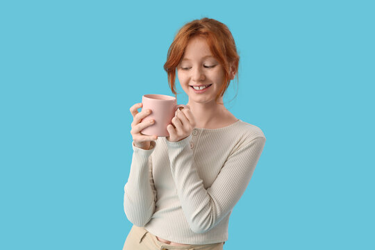 Teenage redhead girl with cup of sweet cocoa on blue background