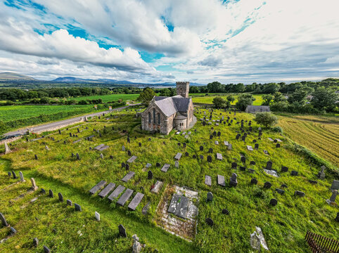 Aerial view of St Nidan's Church and its historic graveyard surrounded by lush green fields and distant mountains under a cloudy sky in Llanfairpwllgwyngyll, Wales, United Kingdom.
