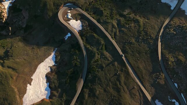Beartooth Highway in Montana curves dramatically through rugged alpine landscape with melting snow patches. Aerial view captures cars navigating the serpentine road at golden hour.