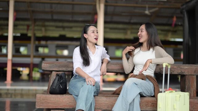Two women sitting on a bench at a train station. One of them is holding a suitcase. Scene is relaxed and casual
