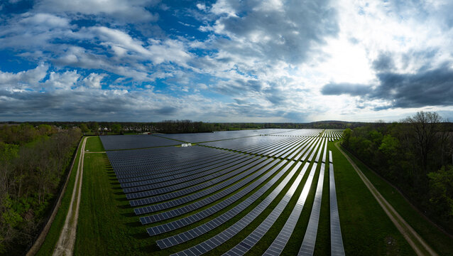 Aerial view of a large solar farm with rows of photovoltaic panels surrounded by lush green trees under a cloudy blue sky Indianapolis, Indiana, United States.