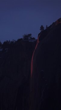Vertical Screen: Yosemite National Park Firefall phenomenon captures Horsetail Fall glowing orange and red against dark granite cliff face during twilight with silhouetted pine trees on ridge