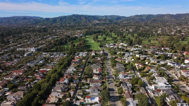 Aerial view los angeles suburb meeting green fairways and distant hills, tree lined corridor guides eye toward golf course, residential roofs and pools pepper landscape under clear sky,