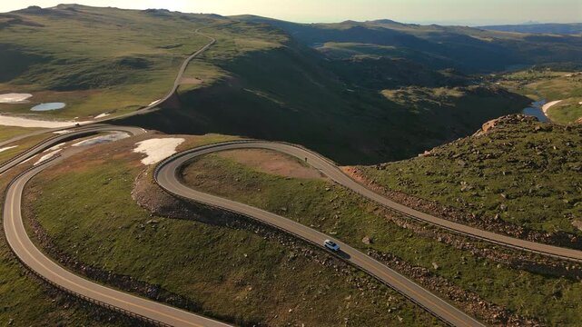 Aerial footage captures a white car navigating the serpentine curves of Beartooth Highway in Montana. Golden hour light illuminates the alpine tundra and distant mountain valleys.