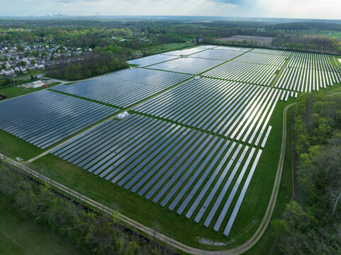 Aerial view of a large solar farm with rows of photovoltaic panels reflecting sunlight near a residential neighborhood and green forests Indianapolis, Indiana, United States.