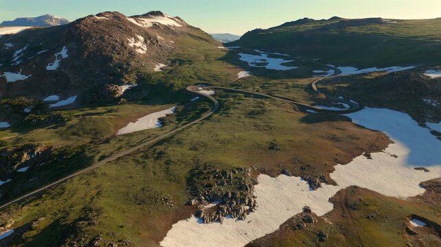 Beartooth Highway in Montana curves through stunning alpine terrain with melting snow patches and lush green meadows. Golden hour light illuminates rocky peaks and winding asphalt roads.