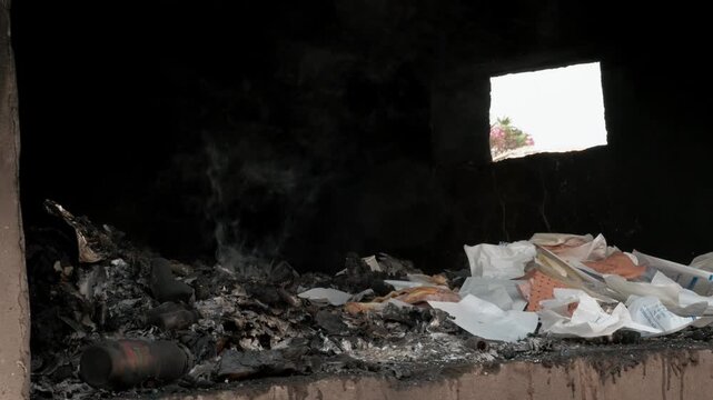 Dark interior of a structure filled with smoldering trash and white debris in Yaounde, Cameroon, highlighting urban pollution issues. Wide shot.