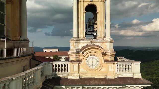 Aerial drone shot tilting up the baroque yellow bell tower and clock face of the Basilica of Superga near Turin, revealing dramatic clouds and the wooded hills of Piedmont.