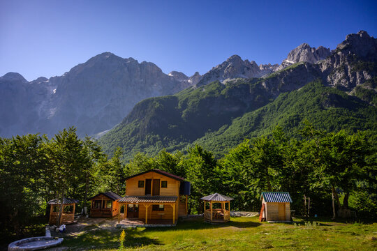 A cluster of rustic wooden guesthouses and small cabins in a sunlit grassy clearing, with towering, rocky mountains forming a dramatic backdrop in Valbona Valley, Albania.