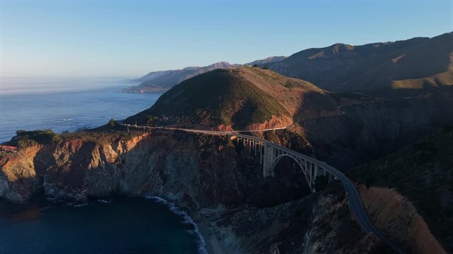 Aerial coastal sunrise over winding bridge cliffs, rugged pacific shoreline bathed in golden light, cinematic drone sweep revealing remote highway hugging canyon, gentle waves lapping rocky