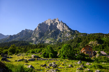 Fototapeta premium A brown lodge is nestled in a rocky green meadow, dominated by a massive, imposing mountain peak and surrounding forests under a bright blue sky in Valbona Valley, Albania.