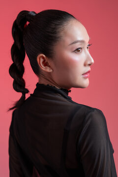 stunning young East Asian woman showing her side profile, featuring a sleek high braided ponytail and flawless glowing makeup. She is wearing a black sheer turtleneck top against a vibrant pink studio