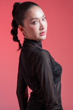 stunning young East Asian woman showing her side profile, featuring a sleek high braided ponytail and flawless glowing makeup. She is wearing a black sheer turtleneck top against a vibrant pink studio