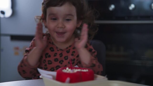Little girl blows out birthday candle with delight. She wears a patterned top and claps hands. Red cake sits on white plate in kitchen. Kitchen oven visible behind her