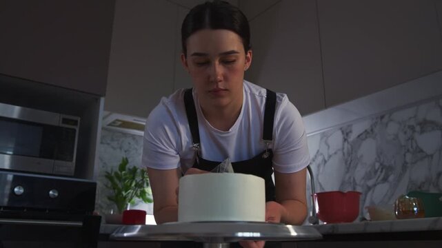 Young woman focuses intently on cake decorating. She holds a piping bag, ready to add details. Kitchen background features marble backsplash and appliances. Her expression shows concentration and care