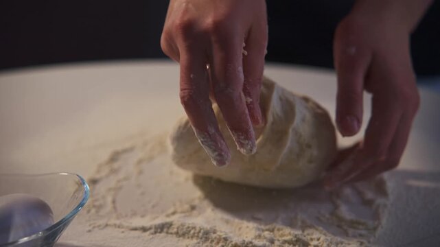 Hands knead dough on a flour-dusted surface. Soft lighting highlights the tactile process. No visible facial expression or emotion. Ideal for cooking, baking, or food preparation themes