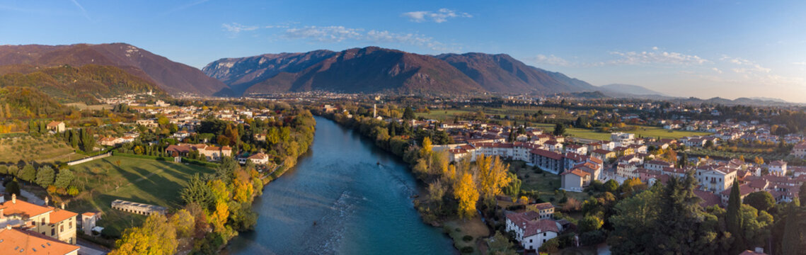 Aerial view of the Brenta river flowing through the historic town of Bassano del Grappa with autumn foliage and mountains Bassano del Grappa, Veneto, Italy.
