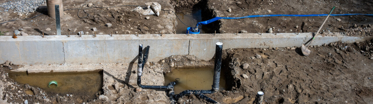 View from above of pipes installed in dirt trenches in foundation of new home build, concrete foundation walls poured, construction work site, pump blue hose pumping out standing rain water
