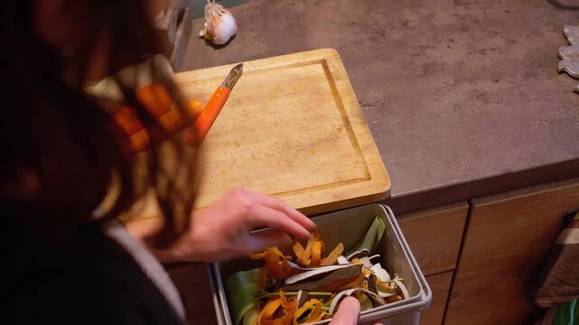 Person collects vegetable scraps like carrot peels in a small grey compost bin on a wooden kitchen counter. Close up view.
