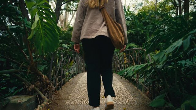 Young woman standing on a small bridge in a lush tropical garden, looking around and taking in the surroundings. Calm and reflective moment in a rich natural environment with dense greenery.