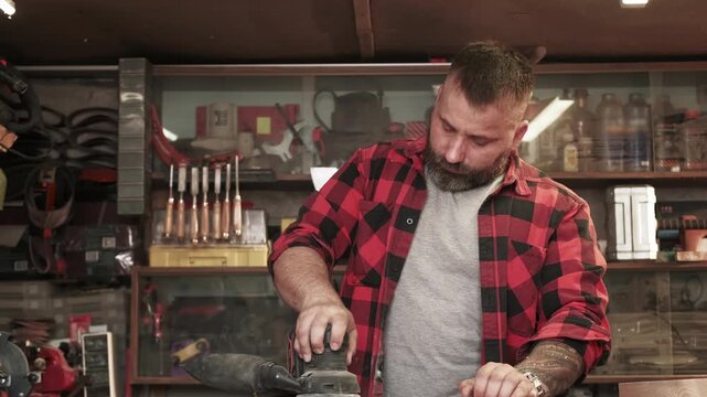 Carpenter occupation. Joinery process. Man smoothing board surface with orbital sander in workshop interior with tools on shelves.
